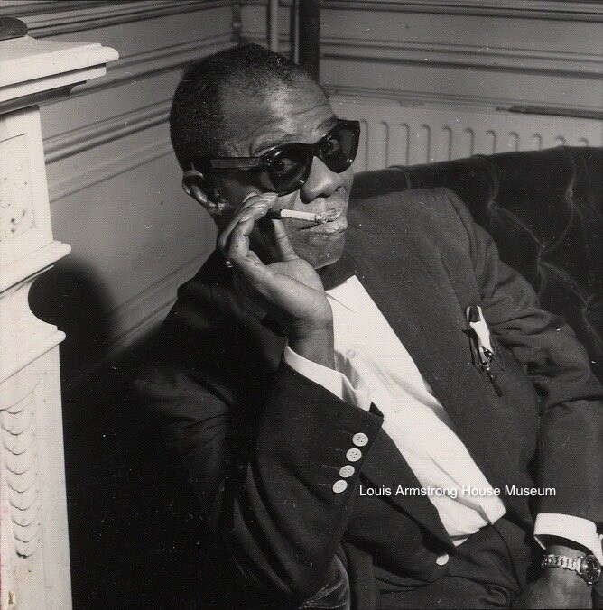 Black and white image of Louis Armstrong during the 1950s. He is wearing sunglasses and smoking a cigarette.