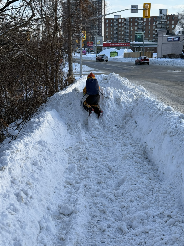 Kid climbs a snow wall on a semi-plowed sidewalk in Ottawa after a snow storm with a big road beside the snow that is fully cleared. 