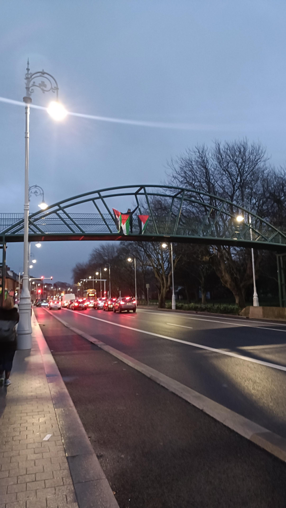 A sound lad stood atop the Fairview pedestrian footbridge waves to the photographer while waving a Palestinian flag. Two additional Palestinian flags hang from the railing of the footbridge. It is dusk.