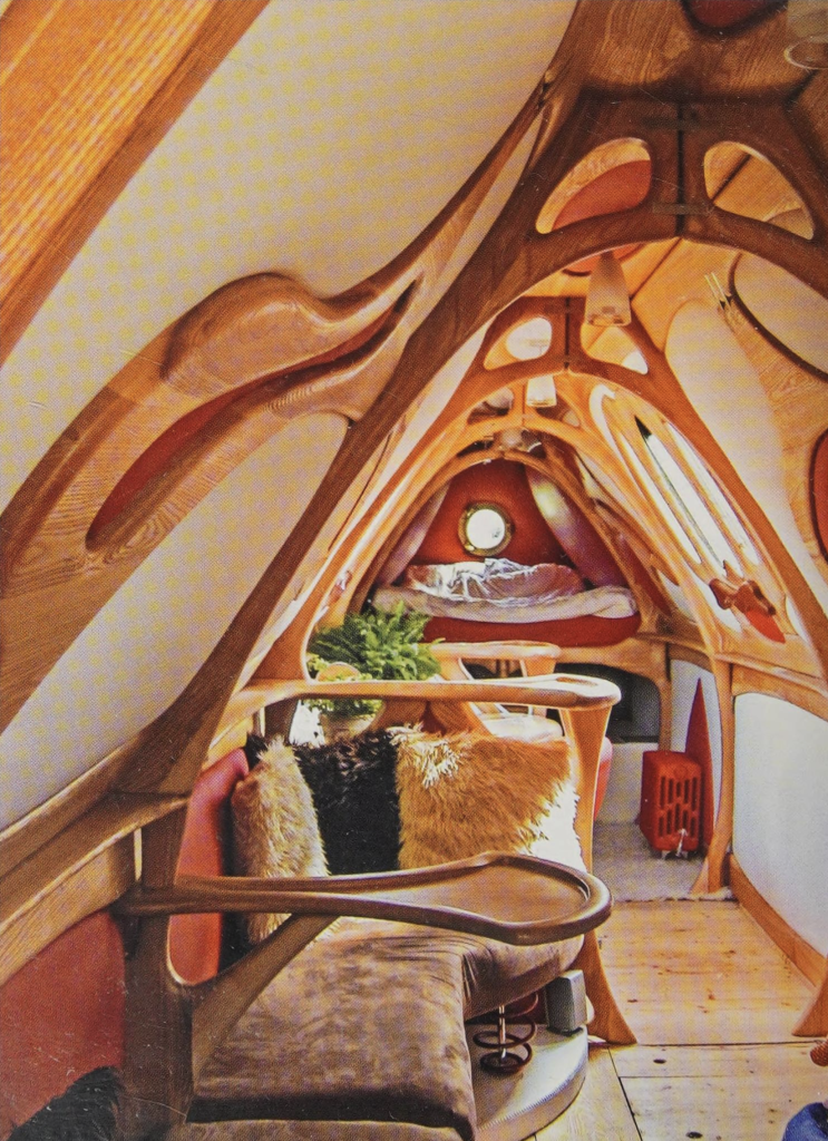 A color photo of the hand-built interior of a small canal boat. The structure and furniture is all made of wood shaped into flowing, organic forms. Welcome.