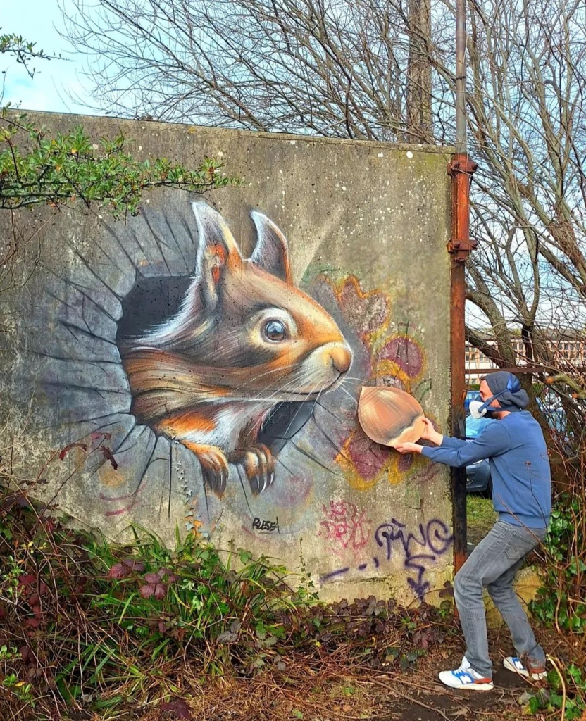 Streetartwall. A funny 3D mural of a giant squirrel was sprayed/painted on an old concrete street wall. The head of a man-sized squirrel peeks out of a (painted) tree hole in search of food. The artist stands in front of the wall and holds a hazelnut (made of cardboard) towards the squirrel, probably out of fear of being eaten himself. Nice effect on what used to be a really ugly concrete wall.