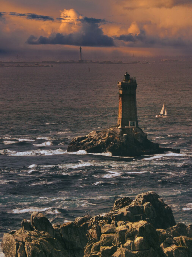 The image depicts a lighthouse standing on a rocky island, surrounded by turbulent ocean waters. In the background, another lighthouse is visible on the horizon under a dramatic sunset sky filled with clouds. A sailboat navigates through the waves nearby.