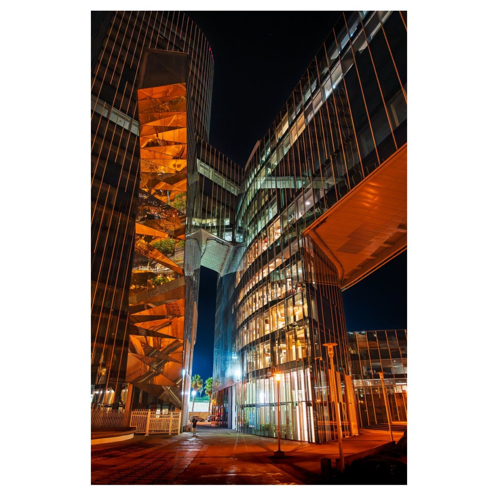 Hochmoderne, gläserne Wolkenkratzer stehen in einer urbanen Umgebung bei Nacht. Säulen aus Licht durchbrechen die Dunkelheit und erhellen die fassadengestützten Strukturen. Eine spannende architektonische Verbindung zieht sich zwischen den Gebäuden, während der Gehweg dezent mit Laternen beleuchtet wird.
