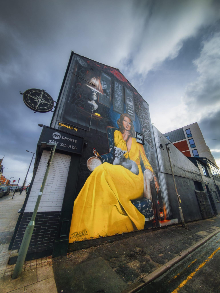 A striking mural on the side of a building in Preston, depicting a woman in a flowing yellow dress seated in a decorative chair. She has a calm expression and holds a glass, while a small lamb rests on her lap. The background includes various artistic elements, such as a lamp and framed images, contributing to a cosy atmosphere. The building itself is painted in darker tones, which contrasts with the vibrant yellow of the dress. In the foreground, street signs and a lamp post are visible, while the sky above is overcast, adding depth to the scene.
