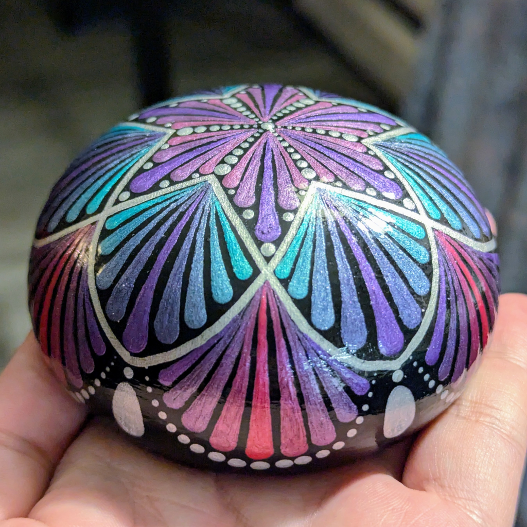 A photo of a dot painting on a round plaster stone. the design consists of a six-pointed star shape filled with radiating teardrop shapes in metallic colours of purple, pink, and turquoise. The star in the centre is circled with scallop shapes filled with the same teardrop starburst patterns. It's punctuated with metallic silver lines and dots.