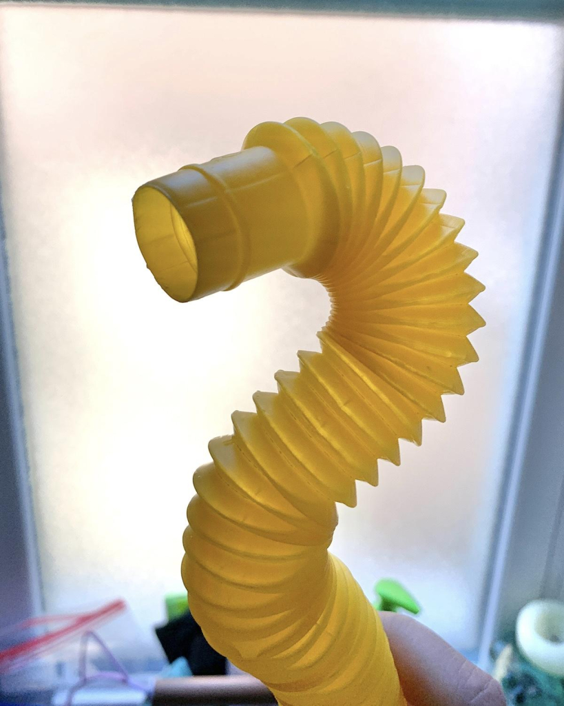 Low quality photo of a kid’s yellow bendy tube toy shaped roughly into an “S” shape with the open end angled toward the viewer, being held by my hand, against my bathroom window with clutter out of focus behind