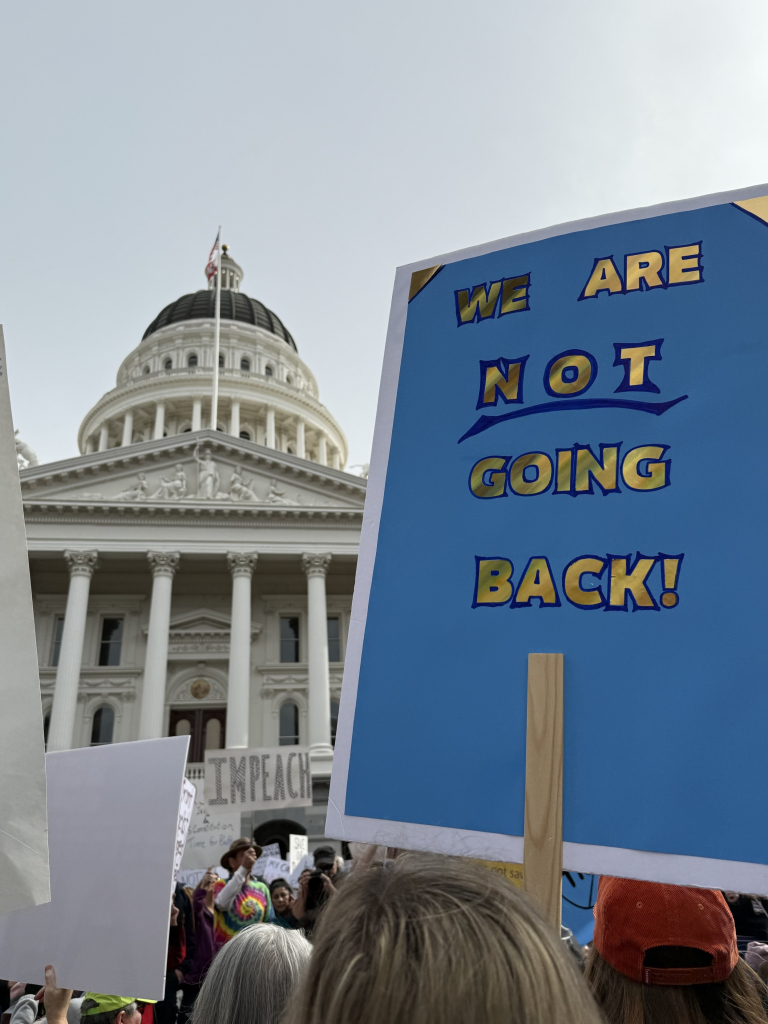 A protest scene in front of the California State Capitol building with people holding signs. One prominent sign reads "WE ARE NOT GOING BACK!" The crowd includes individuals of diverse appearances, some with additional signs advocating for impeachment. The building features a large dome and flags