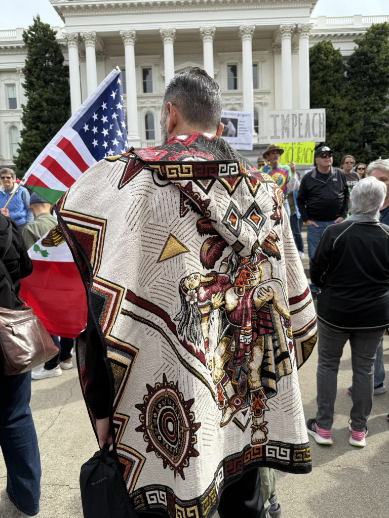 A person in a colorful, patterned poncho stands facing away, holding a black bag. The background features a group of people, an American flag, and a Mexican flag, with a government building visible. Numerous protest signs are in the background, including a sign saying "IMPEACH.”