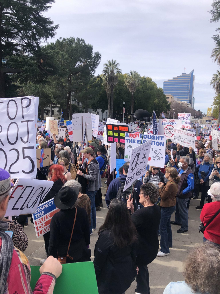 A few thousand people showed up to protest the ongoing coup. This picture shows lots of people with signs and a building in the background in Sacramento, the California Capitol. 