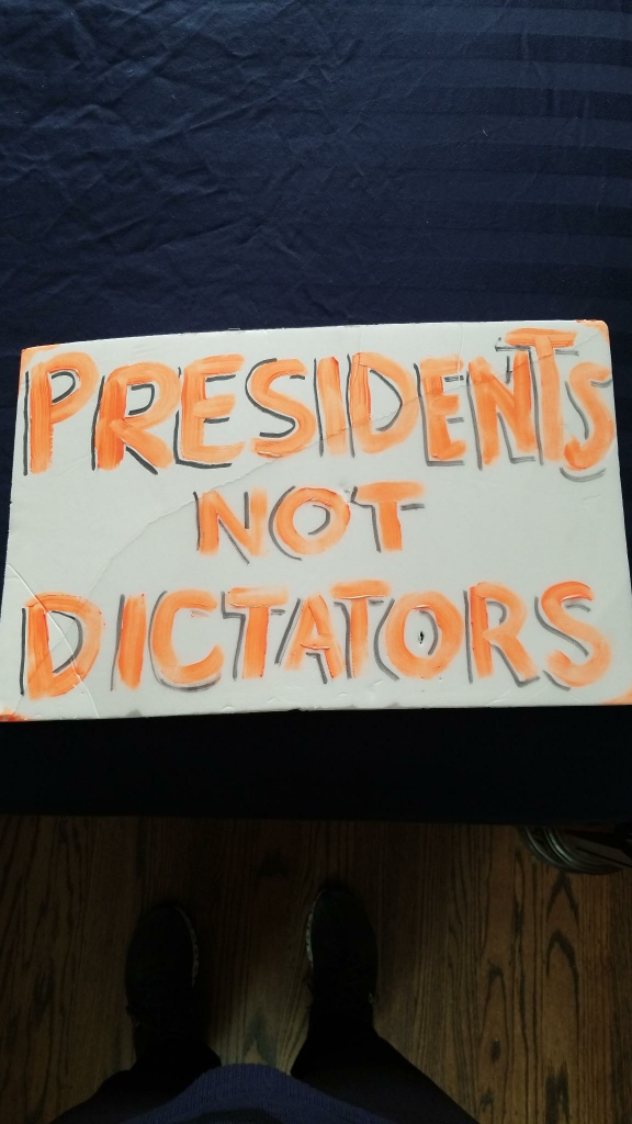 White sign that says "PRESIDENTS NOT DICTATORS" in orange neon with black marker partially outlining the letters