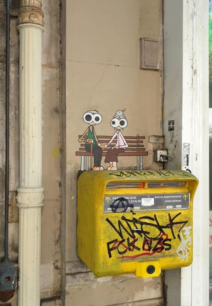 Streetartwall. On a narrow exterior wall between two stores, a small self-painted paste-up with an elderly couple in comic style has been stuck somewhat hidden. The two seniors are sitting on a wooden bench directly above a yellow letterbox and holding hands. The figures both have googly eyes of different sizes and are wearing old-fashioned clothes; he is holding a walking stick. They both look at the viewer. A wonderful idea, because nobody expects to see art in this niche between a bar and a pharmacy. The photo is a bit older, and paste-ups never last very long. Hopefully it was there for a long time.