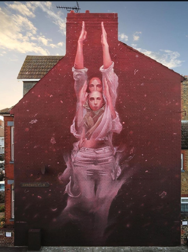 Streetartwall A mural of a woman surrounded by a sea of red was painted on the outside wall of a four-storey modern building. The picture shows a young woman without hair, wearing white jeans and a white blouse, who appears to be floating on the wall. She raises her arms in front of her up to her head. Behind her is another person who looks almost exactly the same, with her arms raised high. Everything around her is red with little white snowflakes. The bright colors of the clothes are blurred, as if they are moving or underwater. The snowflakes are interrupted by larger pieces.
Info: The mural has the theme of microplastics and environmental pollution. It shows a person who wants to break out of this cycle. In addition to the snowflakes, small pieces of plastic are floating next to her, detaching from her clothes and polluting the environment.