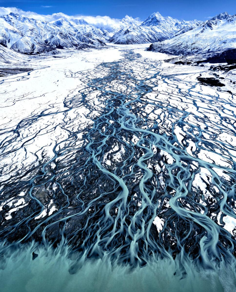 Photography. A breathtaking color photo of a glacial landscape in New Zealand in winter. The aerial view shows the branching glacial Tasman River on the South Island as it flows over the glacial moraine into Lake Pukaki. The branching thin channels of the Tasman, Godley and Cass rivers form a breathtaking tapestry of turquoise hues against the snow-covered landscape. Like a painting, the different shades of turquoise flow in long, gentle waves from the top to the bottom of the picture, where they merge into a narrow strip. Prominent snow-capped mountains (Aoraki/Mount Cook) appear in the distance, majestically framing the terrain. A breathtaking photo in the colors blue/turquoise and white with a certain drama and a skillful perspective that makes the picture appear almost two-dimensional.
Info: The multi-award-winning photo was taken by Stuart Chape. He specializes in aerial photography of landscapes from helicopters and light aircraft, but portraits as well as documentary and social photography are also an important part of his portfolio. The blue coloration of the water in the photo is caused by the refraction of light on fine particles of silt or "glacial flour" in the water, created by glacial erosion.