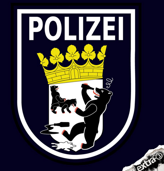 Satirische Darstellung eines Polizei-Wappens, das an das Berliner Polizeiwappen erinnert. Der Berliner Bär, der normalerweise aufrecht und würdevoll steht, ist hier betrunken dargestellt: Er hält eine Flasche in der Hand, taumelt und hat verwirrte Augen. Auf dem Wappen im Inneren sieht man einen weiteren Bären, der scheinbar eine Person mit einem Hund jagt. Auf dem Boden liegen eine zerbrochene Flasche und eine Pfütze. Das Logo der Satire-Sendung 'extra 3' ist in der Ecke zu sehen, was auf eine kritische Auseinandersetzung mit der Polizei hindeutet.