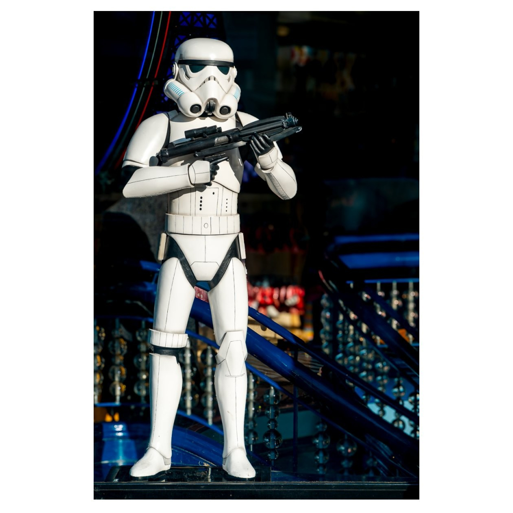 Ein Stormtrooper in weißer Rüstung steht mit einem Gewehr in den Händen. Er ist auf einer Treppe platziert, umgeben von bunten Lichtern und einer urbanen Umgebung, die auf Freizeitaktivitäten hinweist. Das Bild vermittelt ein Gefühl von Action und Spannung.