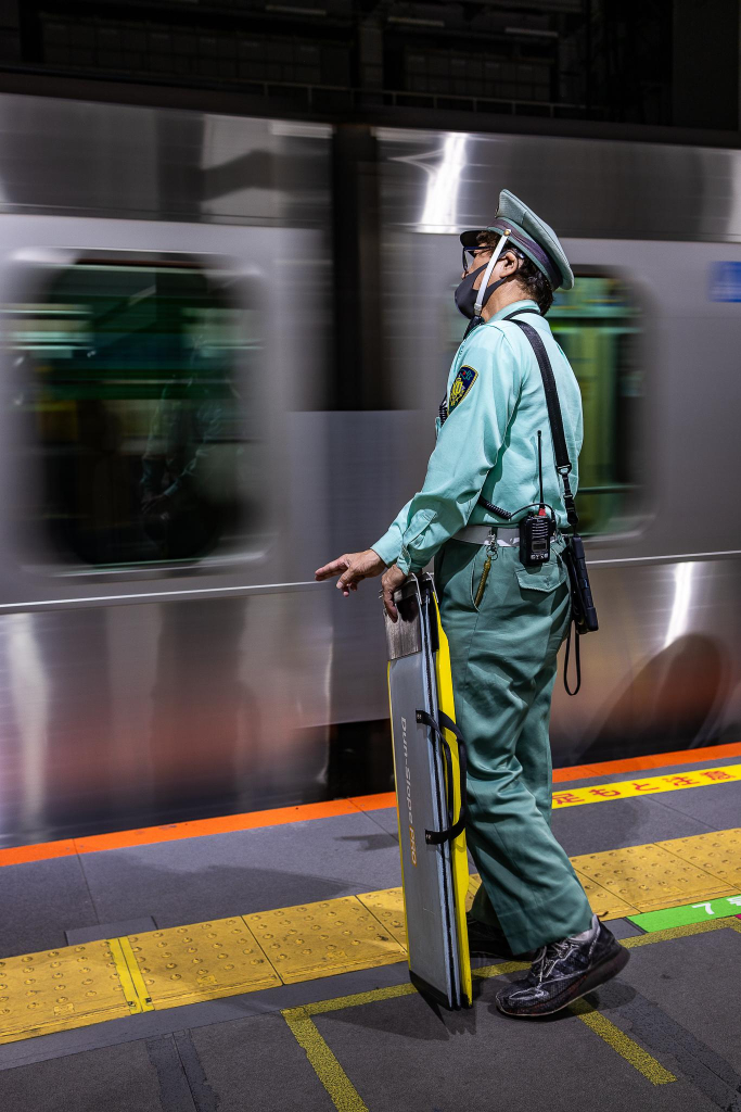 Vertical shot of a Japanese railway officer in a light green uniform and a cap is waiting for a train to stop, with a view to set a mobile bridge to help a disabled passenger get on board, at the Shibuya train station in Tokyo at night. The officer is contrasting nicely with the background made of a platform covered by colorful yellow and orange warning stripes and the blurry grey train entering the station. The train is slightly brushed by the slow motion picture whereas the officer is sharp. 