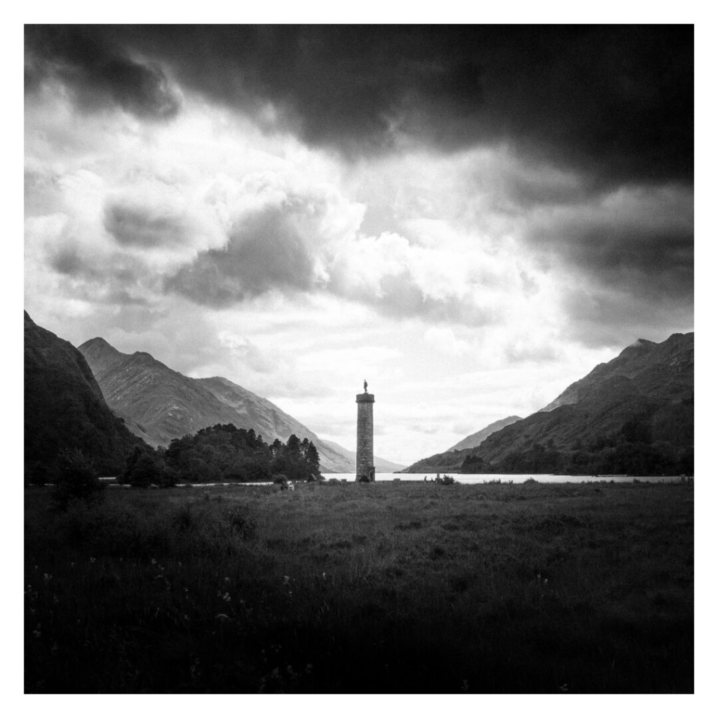 This black-and-white photograph, framed with a white border, shows the Glenfinnan Monument at the shore of Loch Shiel, surrounded by mountains. The monument, positioned centrally, creates a focal point amidst the rugged terrain. A body of water in the background enhances the depth of the image, while the dark foreground contrasts sharply with the lighter sky, adding to the moody and almost cinematic atmosphere. The sky is filled with clouds, and the surrounding landscape consists of rolling hills and vegetation.