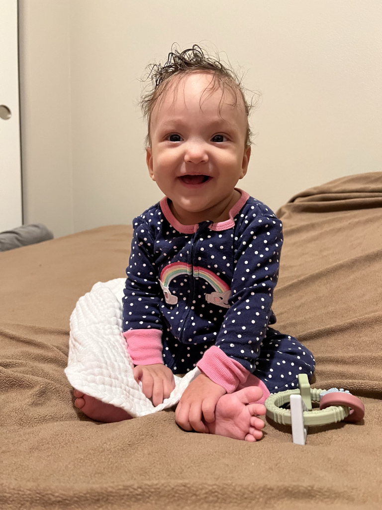 A smiling baby sits on a bed, wearing a navy polka-dot onesie with a rainbow design. The baby has tousled hair and is surrounded by a blanket and a colorful teething toy.