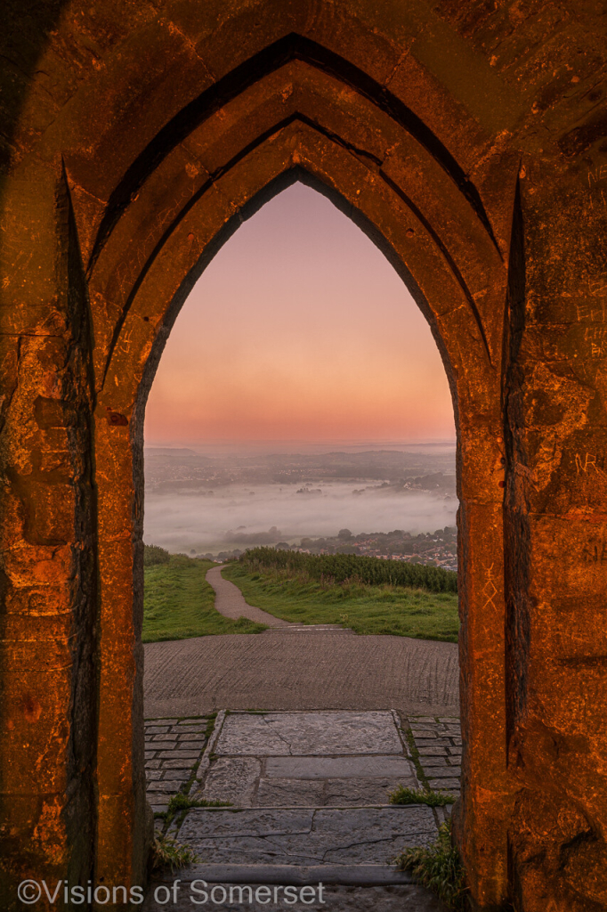 The archway bricks are lit up orange. Path leads down to the town which is covered in mist. Pink and lilac sky above.