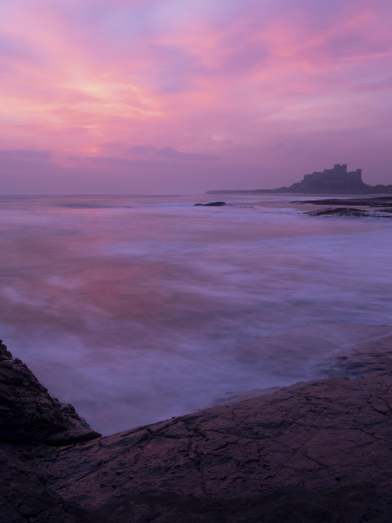 A photo looking across the sea to a castle on a beach at sunrise. The sky is a delicate mix of pink, purple and orange in colour as the sun rises behind cloud on the horizon. The sea has been given a misty appearance by a long exposure, reflecting the colours of the calm areas of its surface. In the foreground the pink hues are reflected back from wet rocks.  