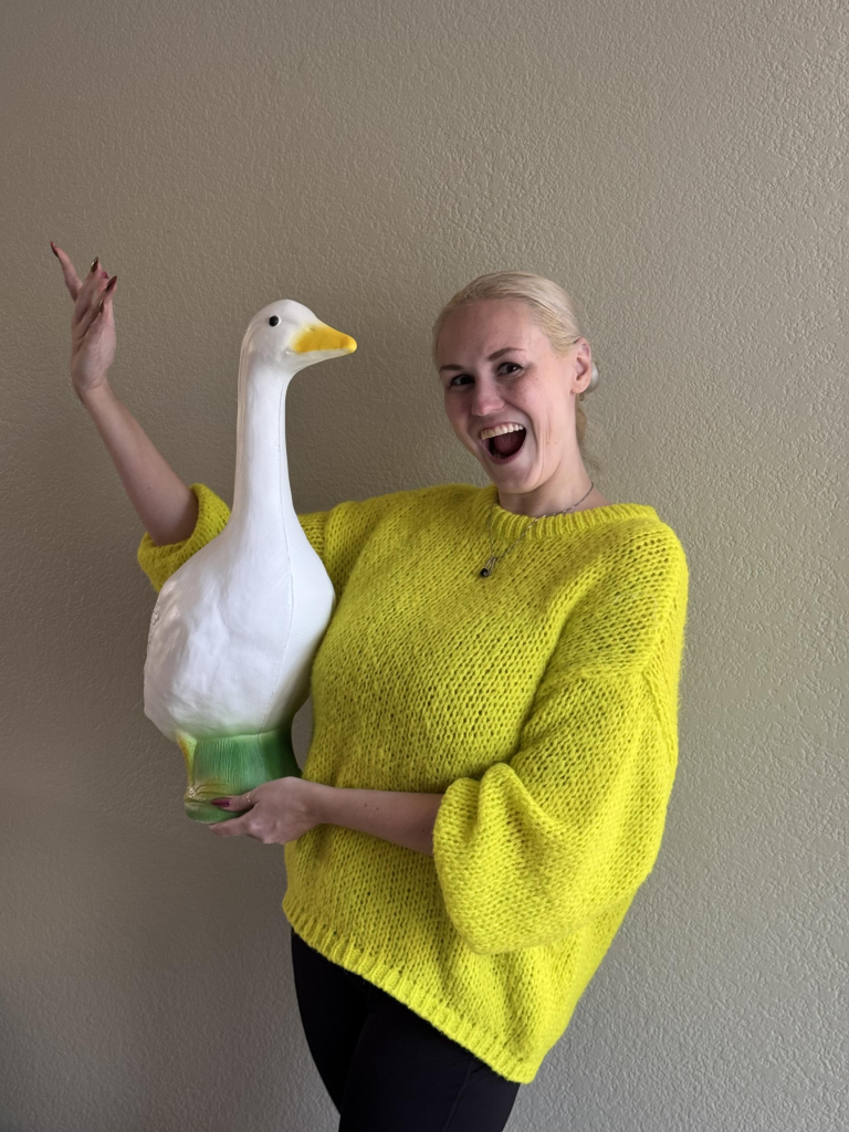 Blonde woman in a bright yellow sweater holding an approximately 2 foot goose statue. She is clearly delighted.