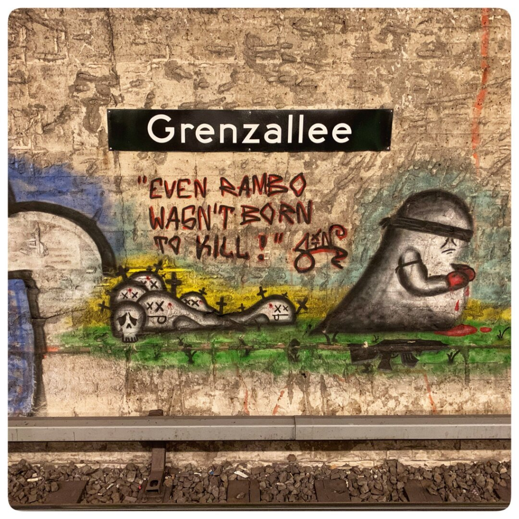 A subway wall with a sign reading "Grenzallee." Below the sign, there is graffiti featuring a character with a bandana and a bloody weapon, alongside skulls and the phrase, "Even Rambo wasn't born to kill!"