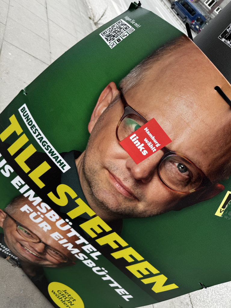 Ein Kampagnenplakat mit einem Mann mit Brille und einem ernsten Ausdruck. Der Text enthält "Bundestagswahl" und "Till Steffen", mit zusätzlichen Details zu den Hamburger Wahlen. Ein roter Aufkleber überlagert einen Teil des Bildes und betont "Hamburg wählt Links“ 