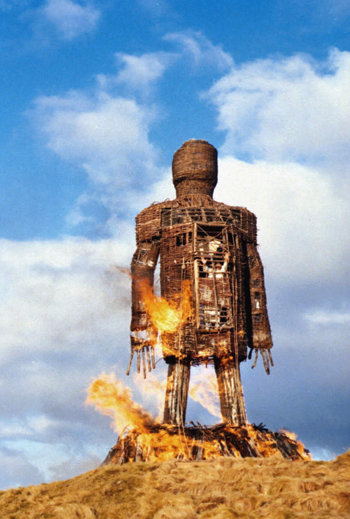 An image of a burning human-looking effigy against a blue sky and puffy white clouds. Inside the effigy is a man in a white robe screaming.