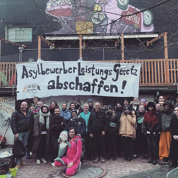 Foto von ca. 30-40 Menschen unter einem Balkongeländer auf dem Hof des UJZ Korn in Hannover. Am Geländer hängt ein Transpi mit dem Text: Asylbewerberleistungsgesetz abschaffen!