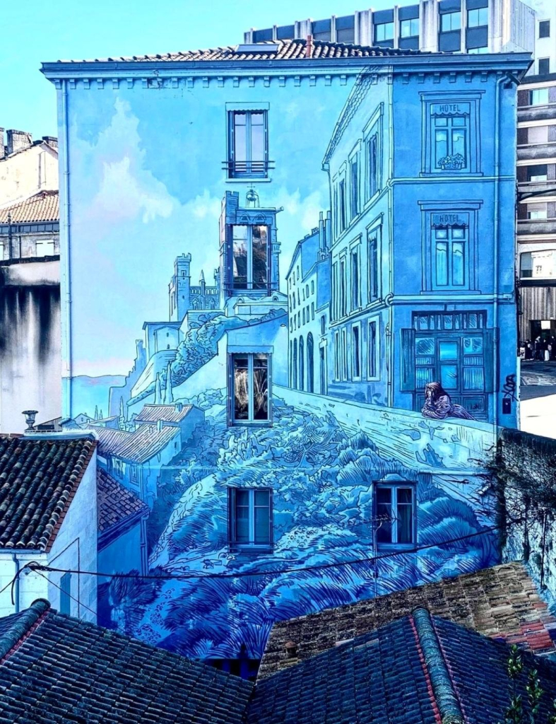 Streetartwall. A fantastic mural based on a comic drawing by Max Cabannes has been installed on the outside wall of a four-storey building on the city wall. The huge mural (120 m²) is painted entirely in shades of blue and shows a girl leaning against the city wall and looking down on the city below. This painted light blue wall connects to the real city wall on the right. The mural shows the view of a town built into the rock on the wall of the house. It incorporates parts of the surroundings, such as roofs and parts of buildings, e.g. the city wall, and continues them in an imaginary view. A wonderfully precise work that extends over several levels and characterizes the cityscape.