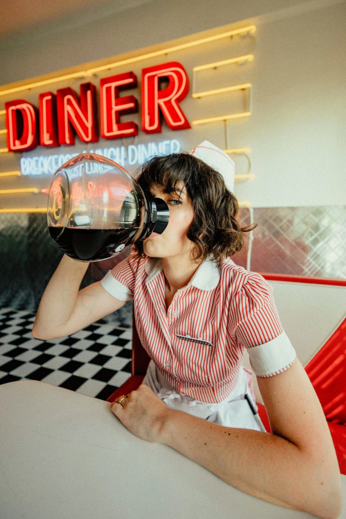 woman sits at a booth in a diner (the word diner behind her in neon), wearing a server's uniform, and drinking directly from an entire glass carafe of coffee