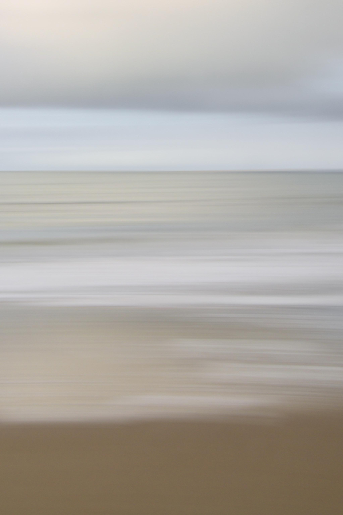 das bild im format 2:3 zeigt eine in bewegung geschossene strand- und meeresszene.
die szenerie ist horizontal geschichtet.
ganz oben ist eine wolkenschicht. darunter ist freier himmel. dann kommt der horizont. darunter in der ferne das weite meer. zum vordergrund hin sieht flache wellen erkennbar. unterhalb der bildhälfte ist der brandungsschaum zu sehen. darunter ist schon nasser sand. und ganz unten als letzter streifen ist nur etwas dunkler und glatter strand zu sehen.
bei der aufnahme wurde die kamera horizontal bewegt, sodass das bild verschwommen ist und eine horizontale bewegungsunschärfe als stilmittel die wahrnehmung der schichten verstärkt. zusätzlich wird eine abstrakte komponente eingebracht.