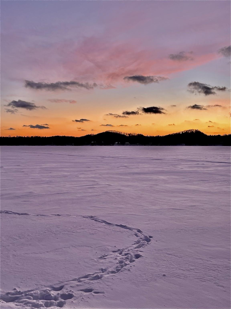 A serene winter landscape featuring a snow-covered frozen lake with Brookie’s footprints in the foreground. The background showcases a colorful sunset with soft pink and orange hues, contrasted by dark silhouettes of distant hills and clouds.