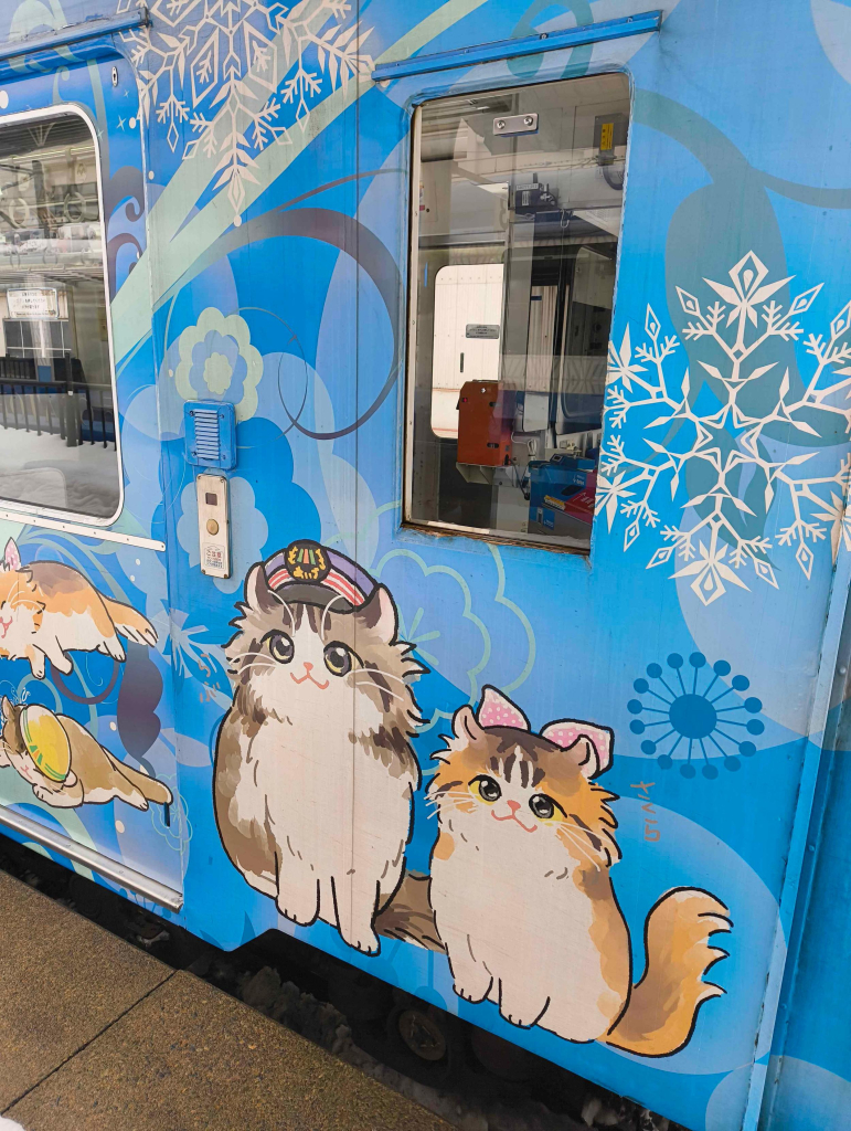 Illustrations of a pair of cats, one of which is wearing a stationmaster’s hat, on a blue diesel railcar train. There are lots of flower and snowflake illustrations in the background of the train livery.