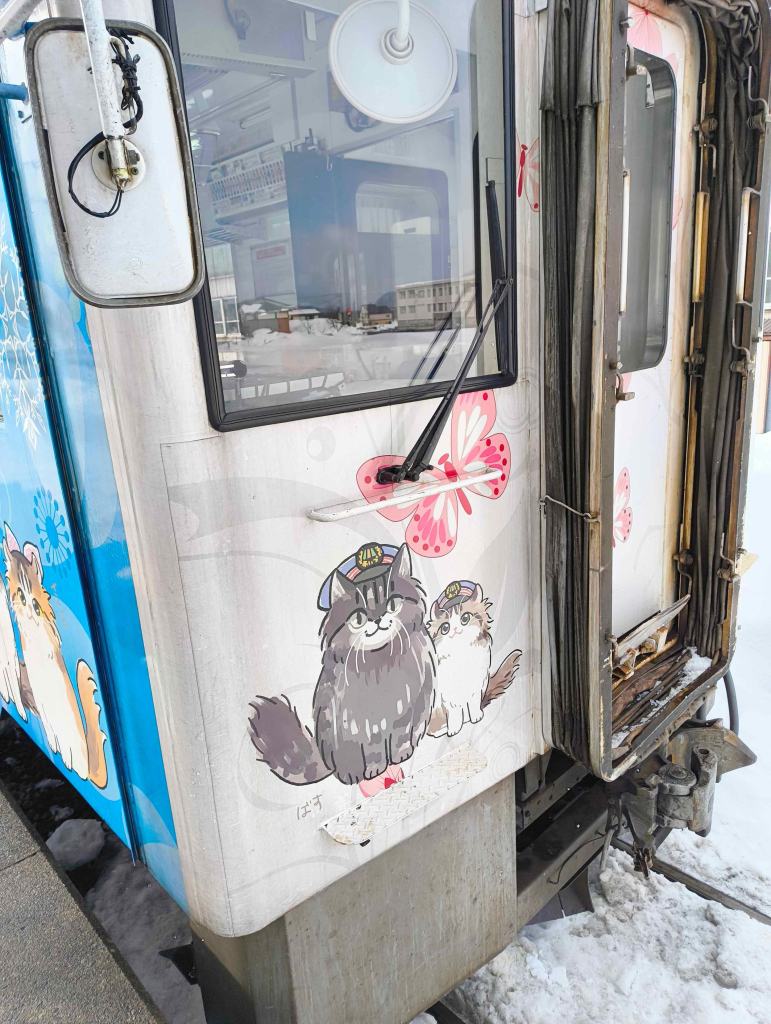 Front of a diesel railcar train at a station platform in the snow. There’s a cartoon of a pair of cats wearing stationmaster hats on the front of the train