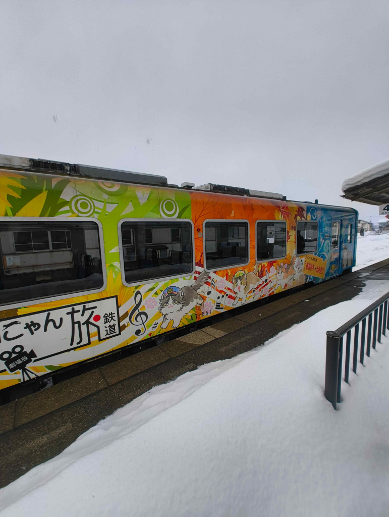 Diesel railcar at a station platform in the snow, wearing blue and yellow livery with cartoon illustrations of the Aizu Railway’s railway cats who live at Ashinomaki Onsen Station