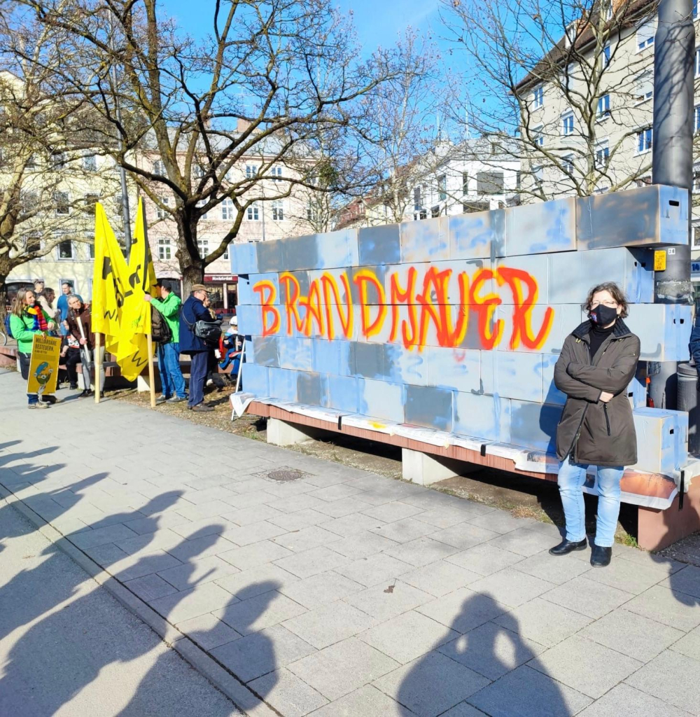 Ich, weiblich, Jeans, olivgrüne Jacke stehe vor einer gebastelten Mauer mit orangener Aufschrift BRANDMAUER. Zusammen mit dem #BündnisGegenRechts in München vor dem Löwenbräukeller.