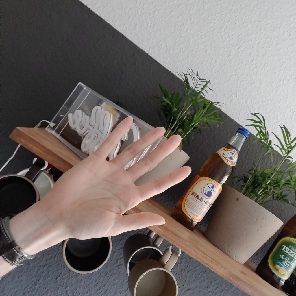 Linke Hand mit einer Armbanduhr darum hoch gestreckt, im Hintergrund steht ne Club Mate, grüne Pflänzchen und ein paar Tassen hängen auch da. Außerdem noch ein Radler.