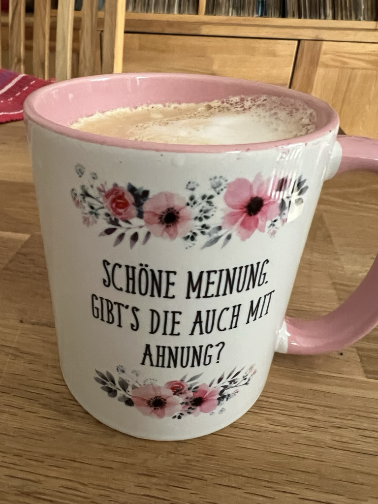 Das Bild zeigt eine Kaffeetasse auf einem Holztisch. Die Tasse ist außen weiß und innen rosa, mit einem rosa Henkel. Verziert ist sie mit einem floralen Muster aus rosa, roten und schwarzen Blüten, das sowohl oben als auch unten auf der Tasse angebracht ist.  Auf der Tasse steht ein humorvoller Spruch in schwarzer Schrift: „Schöne Meinung. Gibt’s die auch mit Ahnung?“  Die Tasse ist gefüllt mit einem Getränk, das wie ein Cappuccino oder Latte Macchiato aussieht – man sieht eine cremige Schaumschicht an der Oberfläche. Im Hintergrund sind Holzmöbel und ein Regal mit Schallplatten oder Büchern zu erkennen. Die Szene wirkt gemütlich und lädt zu einem entspannten Kaffeegenuss ein.