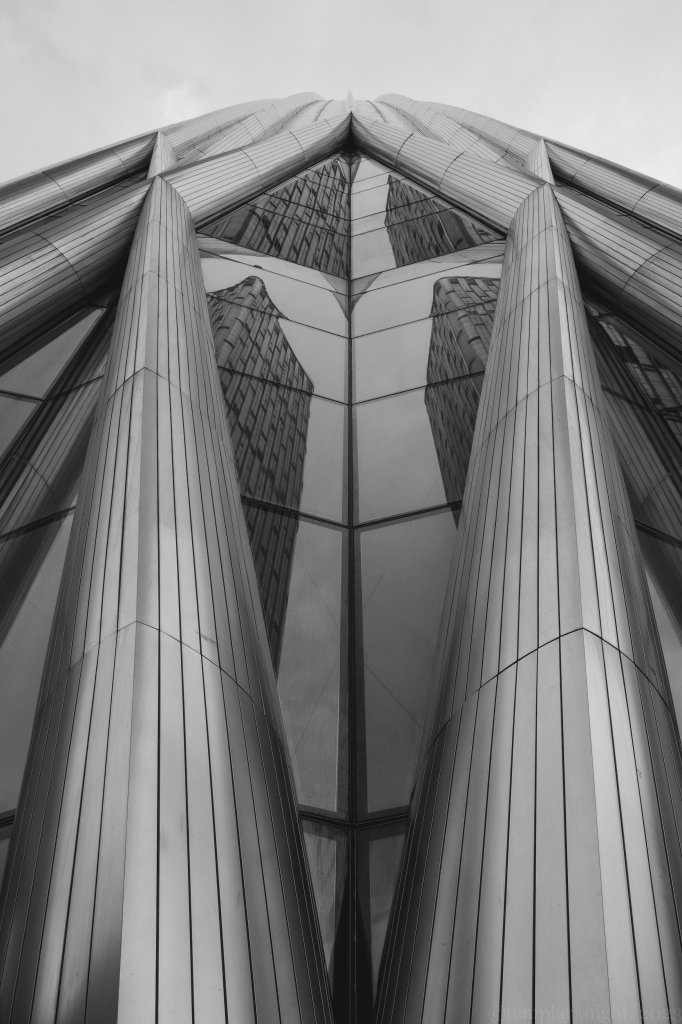 Black and white photo in portrait format of a modern high rise from the perspective on the ground level looking upwards at the glass reflections of the building on a cloudy grey sky. The design and the architecture of the building makes the reflections of the other surrounding office buildings to form a almost a giant human figure.