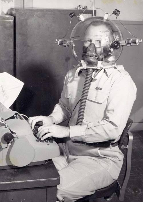 b&w photo of a serviceman of some kind sitting at a typewriter while wearing a breathing apparatus and a weird glass helmet with vacuum tubes and wire around the outside of it