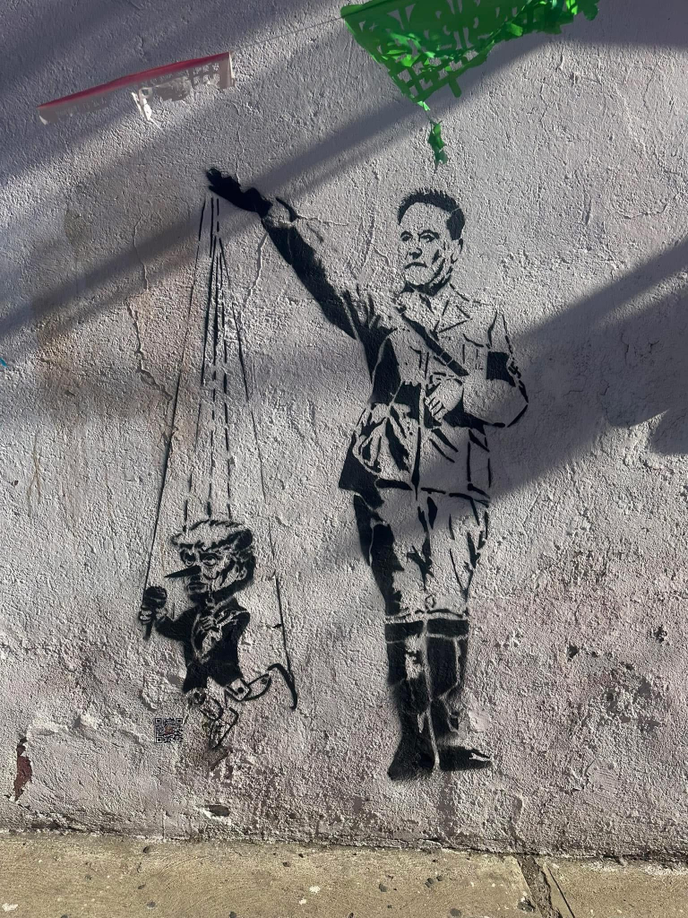 Street art in the style of Banksy on a roughly plastered (stucco?) wall. It depicts Elon Musk in a Nazi uniform using his "Roman Salute" (aka Nazi salute) to puppeteer a little Donald Trump with a Pinocchio nose.