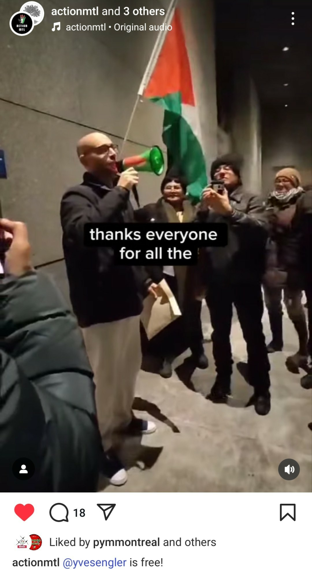 A man with a megaphone speaking around five people, there is a big Palestine flag in the back, it is at night in front of a concrete building