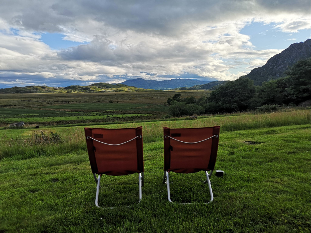 Zwei Campingstühle stehen auf einer grünen Wiese.
Vor den Stühlen breitet such weites flaches Land aus, das mit grünem und braunem Gras bewachsen ist.
In der Ferne erheben sich Felsen und Hügel, auf denen vereinzelte Baume stehen.
Dazwischen sind einzelne von der sonne beschienenene helle Flecke zu sehen