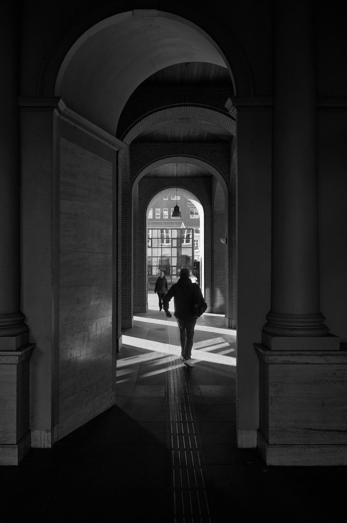 A black and white image of a colonnade with several arches. Two figures are seen walking through the corridor, with light casting geometric patterns on the ground. The background features windows of a building, adding depth to the scene.