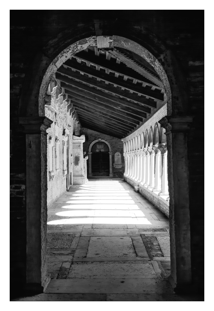 A black and white photo shows a sunlit, arched stone passageway.  The passage is lined with columns on one side, leading to a closed doorway at the far end.  The ceiling is composed of a wooden structure with visible beams.  The stone floor shows the shadows of the arches cast by the sunlight.