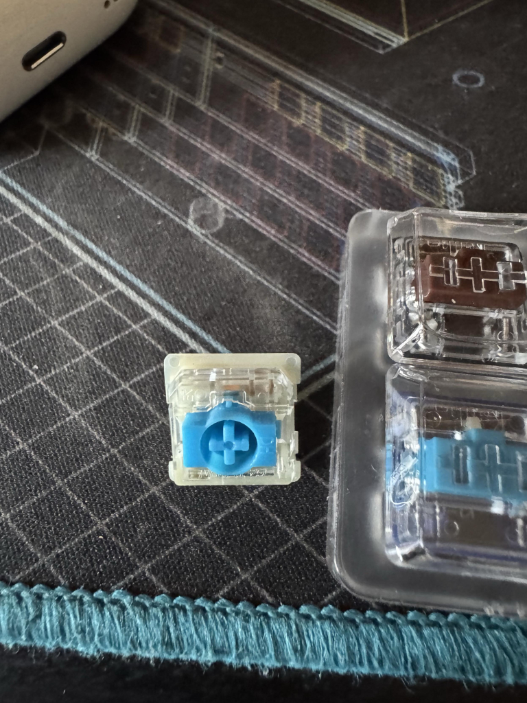 A blue and white keyboard switch without a cap sits facing the camera. The stem pattern is a plus inside a circle 