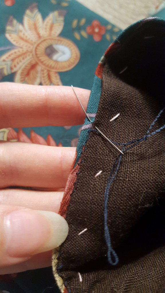 A very close up photo of whipstitching on the inside of the cuff, with a needle and thread poked through the layers, showing how only a tiny little bit of fabric is picked up. The lining edge has been ironed to be a couple of millimetres shorter than the outer layer, thus preventing it from peeking out. The thread is a deep indigo blue while the fabric is teal. The blue thread is barely visible against the dark brown lining.