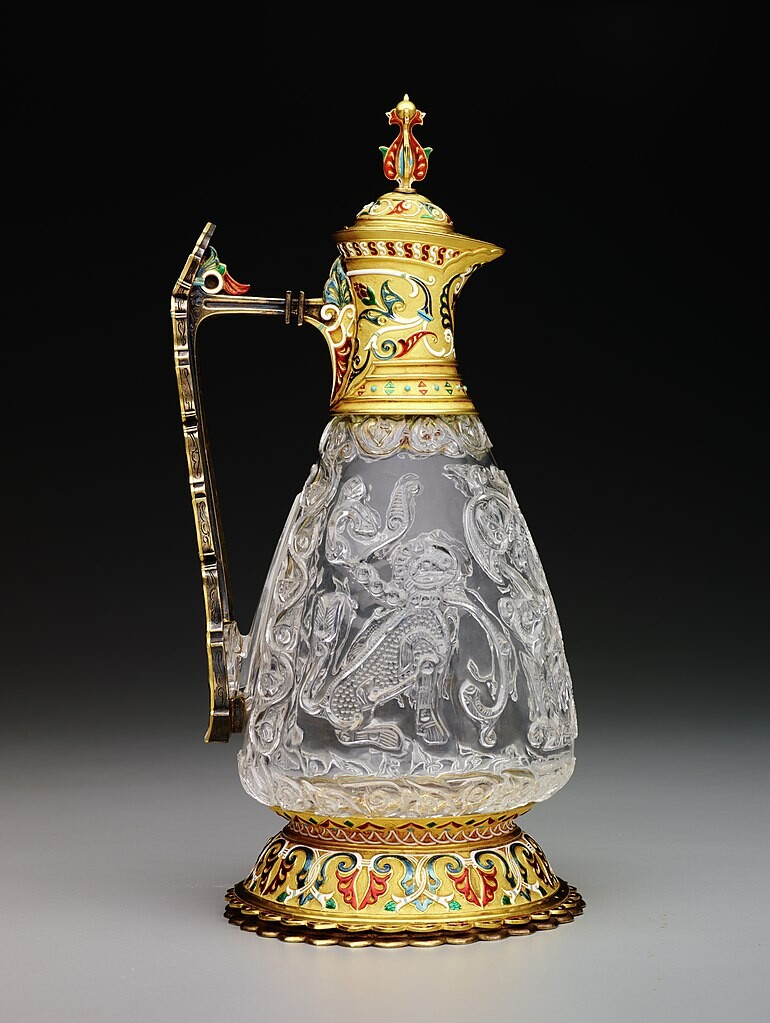 A jug carved from colourless quartz. There are various animals and floral patterns carved into the quartz. The base, handle and lid/spout are made of gold with colourful enamelling.