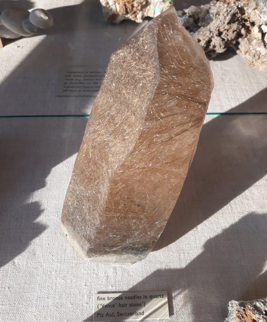 A hexagonal crystal of quartz filled with golden needles of rutile. There are so many rutile needles inside, it almost looks like a mass of golden hair.

Label text: Fine bronze needles in quartz (Venus hair stone). Piz Aul, Switzerland.